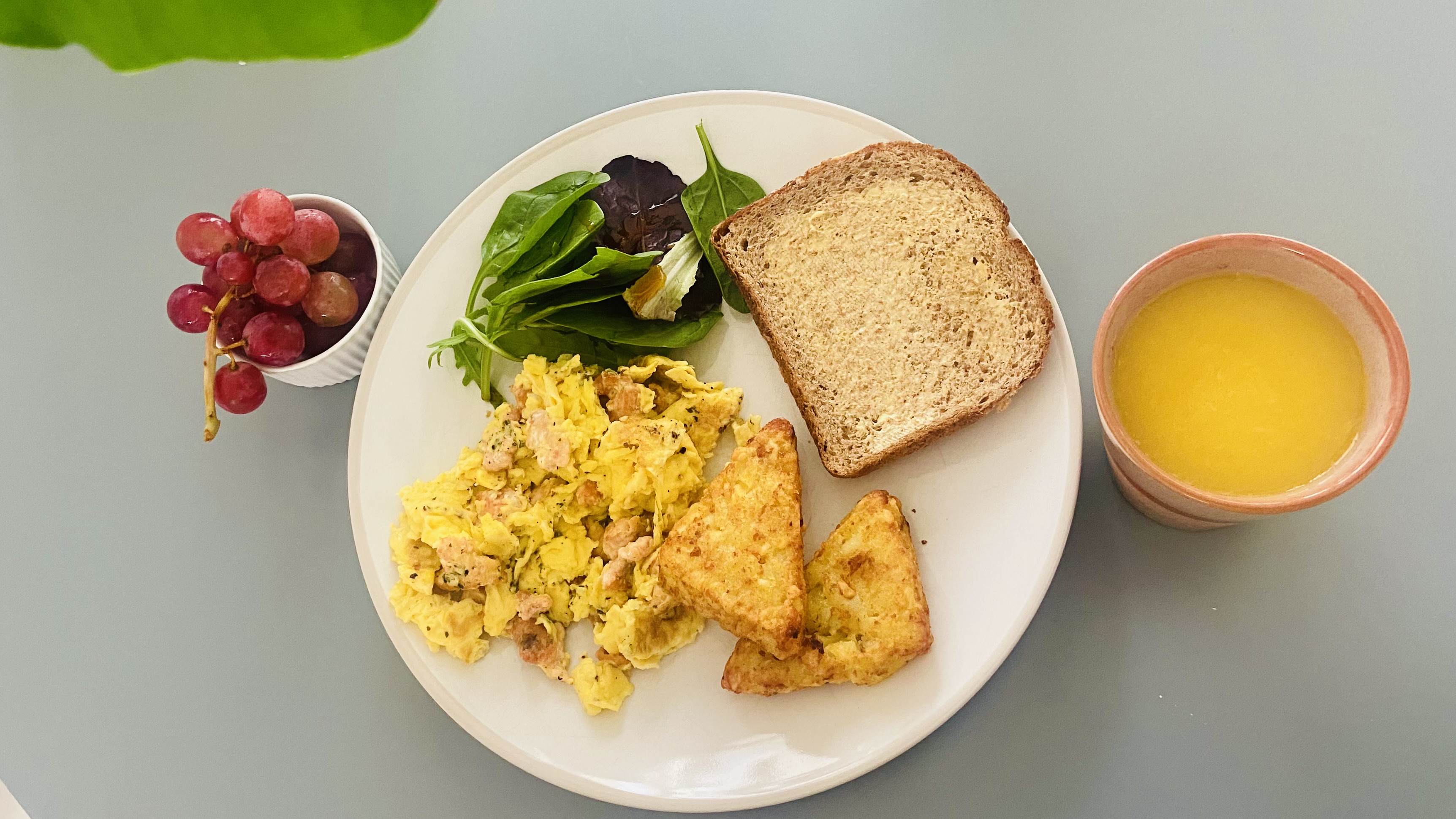 Breakfast of scrambled eggs, hash browns, buttered bread, baby leaves, grapes, and orange juice